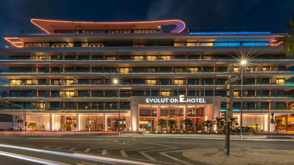 Fachada do Evolution Hotel ao anoitecer, com iluminação linear a contornar a cobertura e letreiro iluminado sobre a entrada.