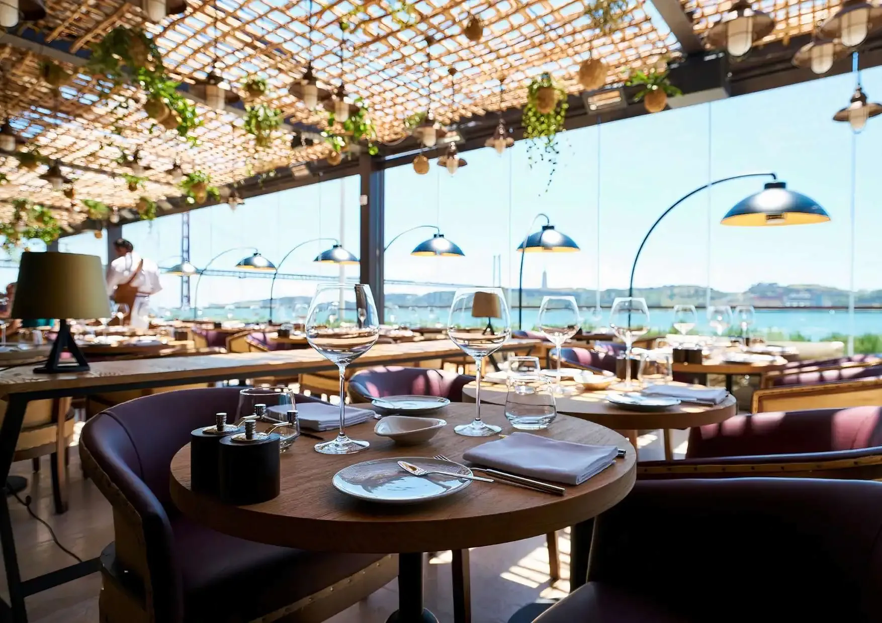 Sala envidraçada sob uma pérgola com plantas suspensas, mesas postas com copos de vinho e o rio ao fundo.
