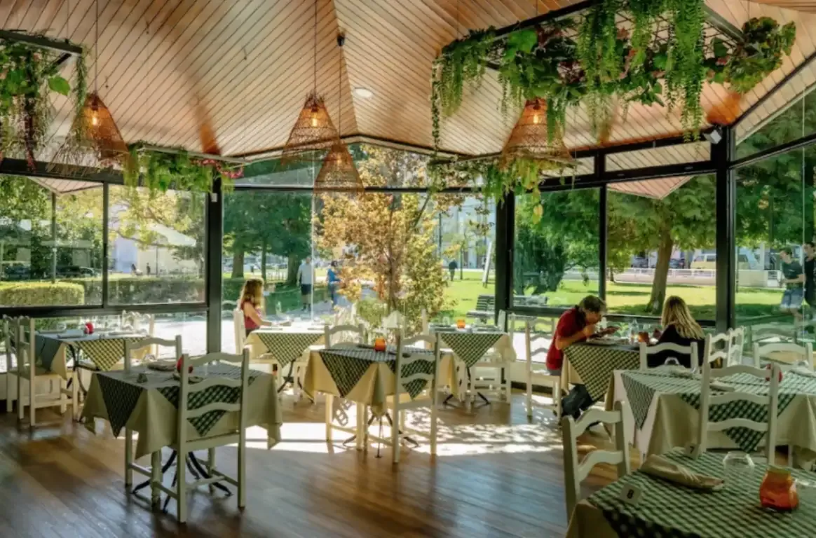 Sala envidraçada virada para um parque, com toalhas aos quadrados verdes, cadeiras de madeira brancas e candeeiros de vime suspensos.