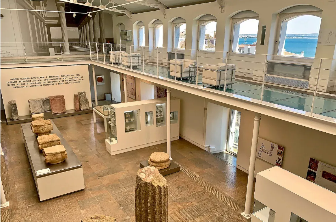 Sala de museu com pedras romanas em expositores, uma inscrição latina na parede e uma galeria envidraçada com vista para o rio.