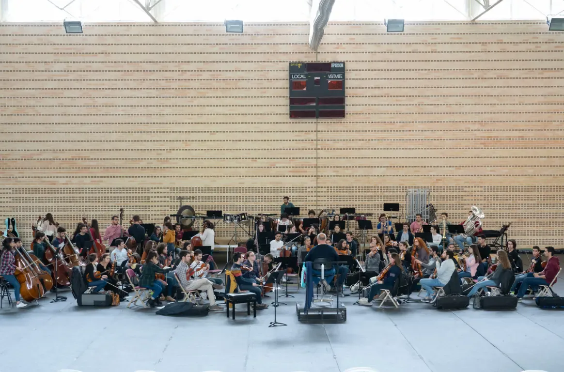 Orquestra em ensaio no recinto de um pavilhão desportivo, com o maestro ao centro e o marcador de jogo na parede de tijolo.