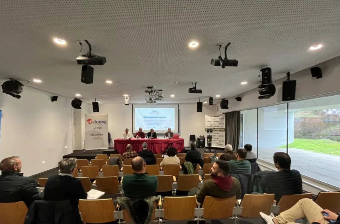 Sala de conferências com público em cadeiras de madeira, uma mesa ao fundo diante de uma tela de projeção e colunas de som no teto.