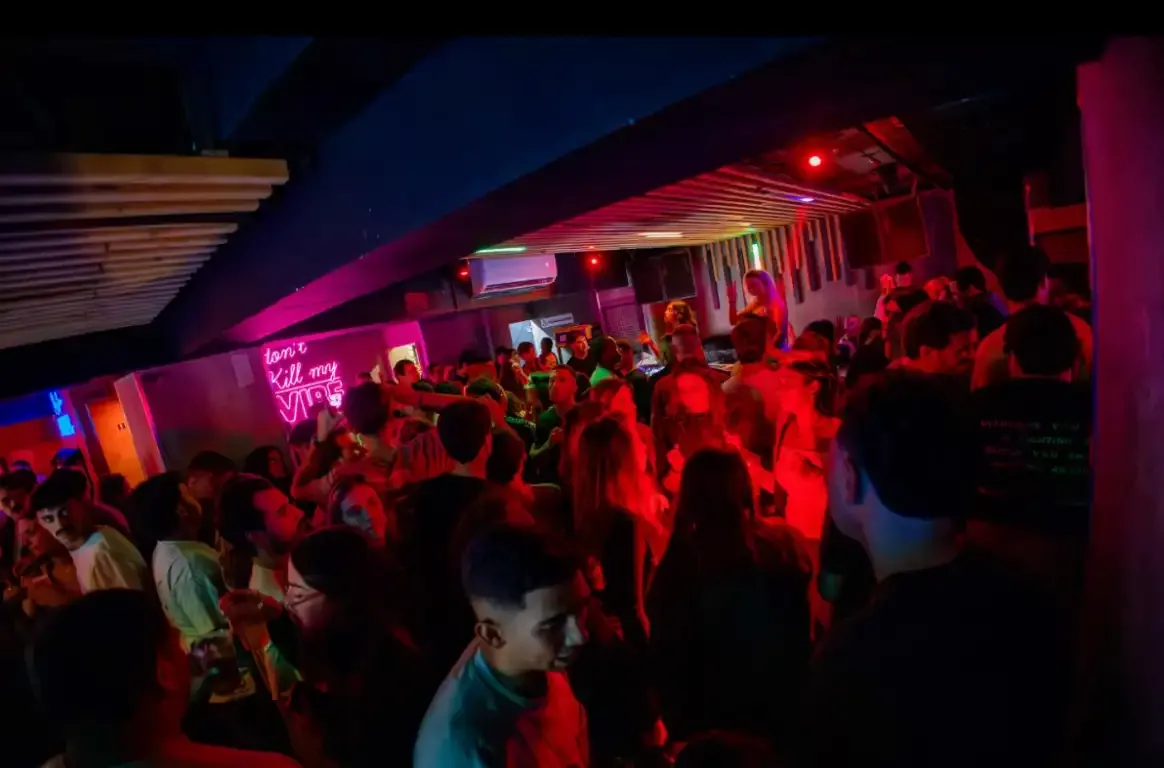 Pista cheia de gente sob luz vermelha, com um letreiro de néon cor-de-rosa na parede ao fundo.