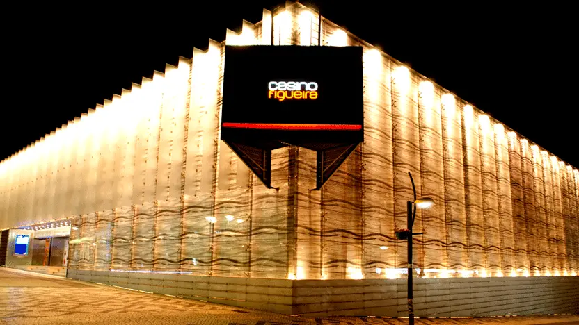 Fachada do Casino Figueira à noite, com a superfície ondulada iluminada e o letreiro central sobre a esquina.