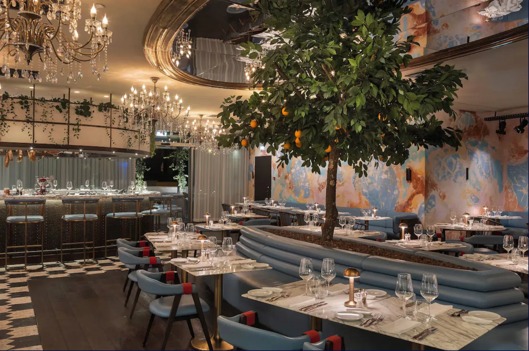 Restaurante com uma árvore ao centro, lustres de cristal, bancos azuis e mesas postas.