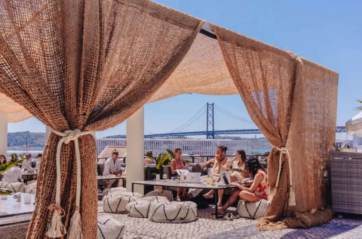 Esplanada num terraço, entre cortinas de juta, com pessoas sentadas em pufes e uma ponte suspensa sobre o rio ao fundo.