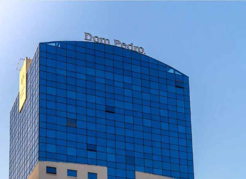 Topo do edifício do Hotel Dom Pedro em dia de sol, com fachada envidraçada azul e letreiro sobre a cobertura.