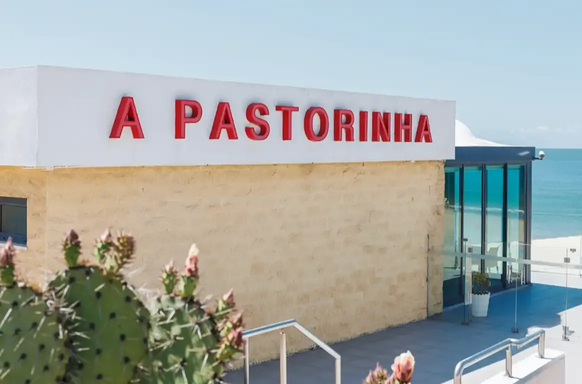 Fachada com o letreiro A PASTORINHA em letras vermelhas, catos em primeiro plano e o mar ao fundo.