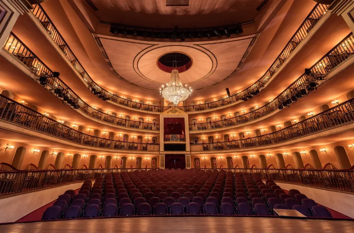 Sala à italiana vista do palco, com plateia de cadeiras roxas, várias ordens de balcões iluminados e um lustre de cristal ao centro.
