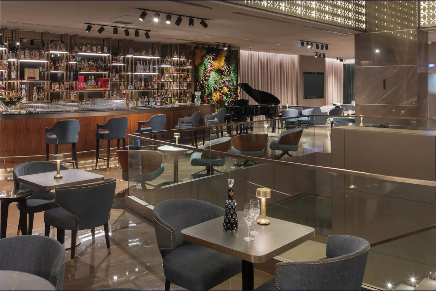 Bar com garrafeira iluminada, piano, mesas com cadeiras cinzentas e guarda de vidro.