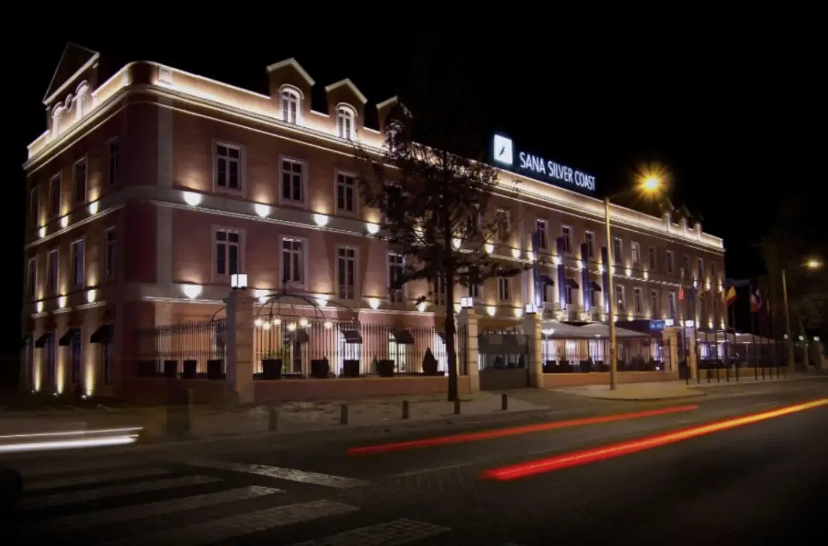 Fachada histórica do hotel à noite, com a iluminação a realçar as janelas e rastos de luz dos carros na rua.