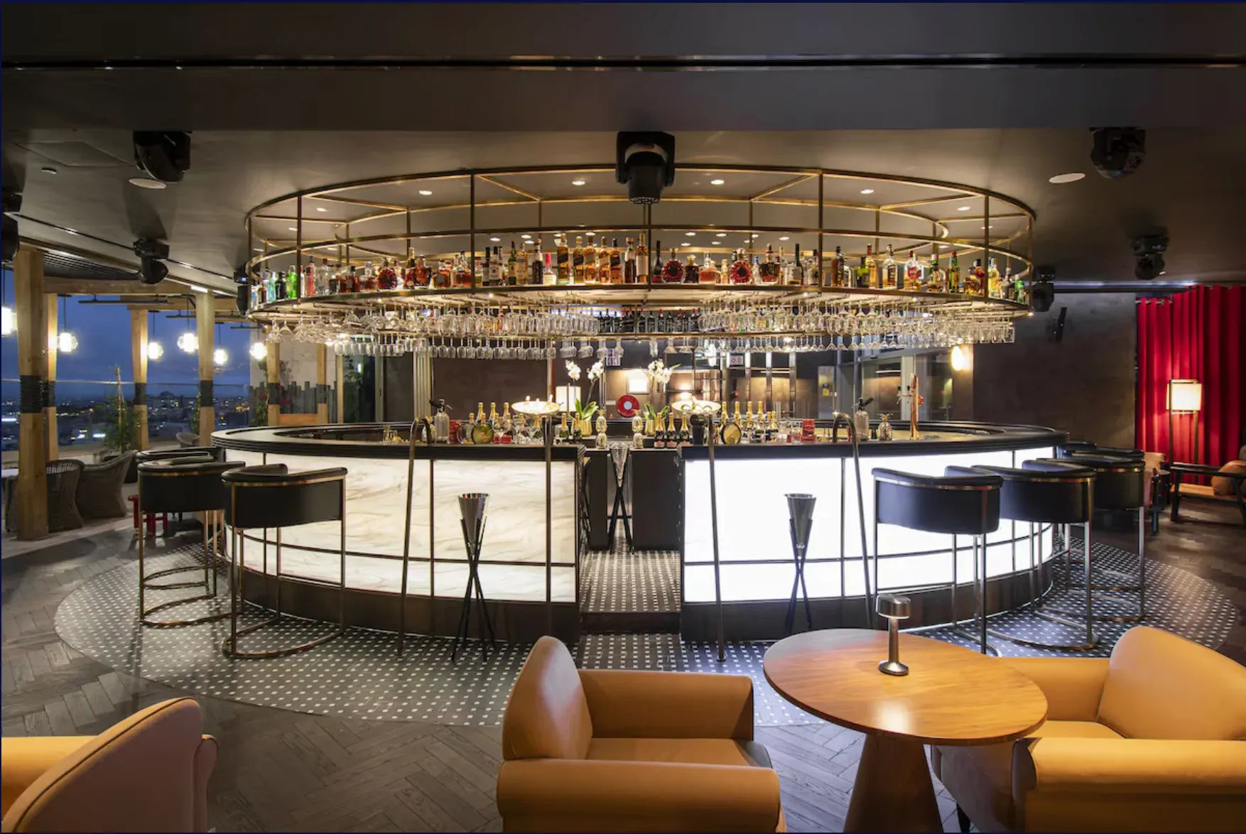 Bar circular com prateleiras de garrafas suspensas, balcão iluminado e poltronas cor de mel.