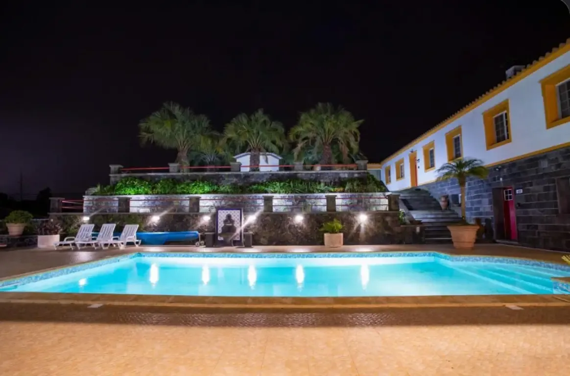 Piscina iluminada à noite, com espreguiçadeiras, palmeiras num terraço de pedra e um edifício caiado de molduras amarelas.