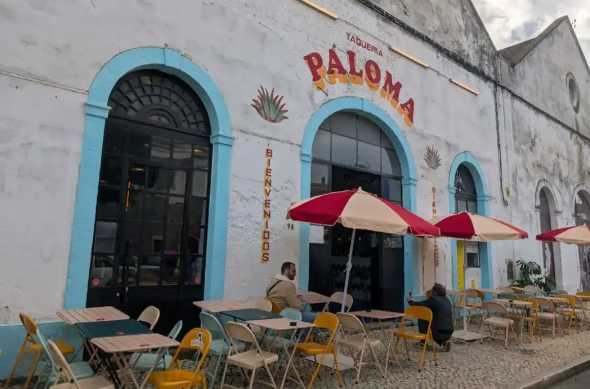 Fachada caiada com arcos azuis e o letreiro TAQUERIA PALOMA, esplanada de cadeiras amarelas e guarda-sóis vermelhos.