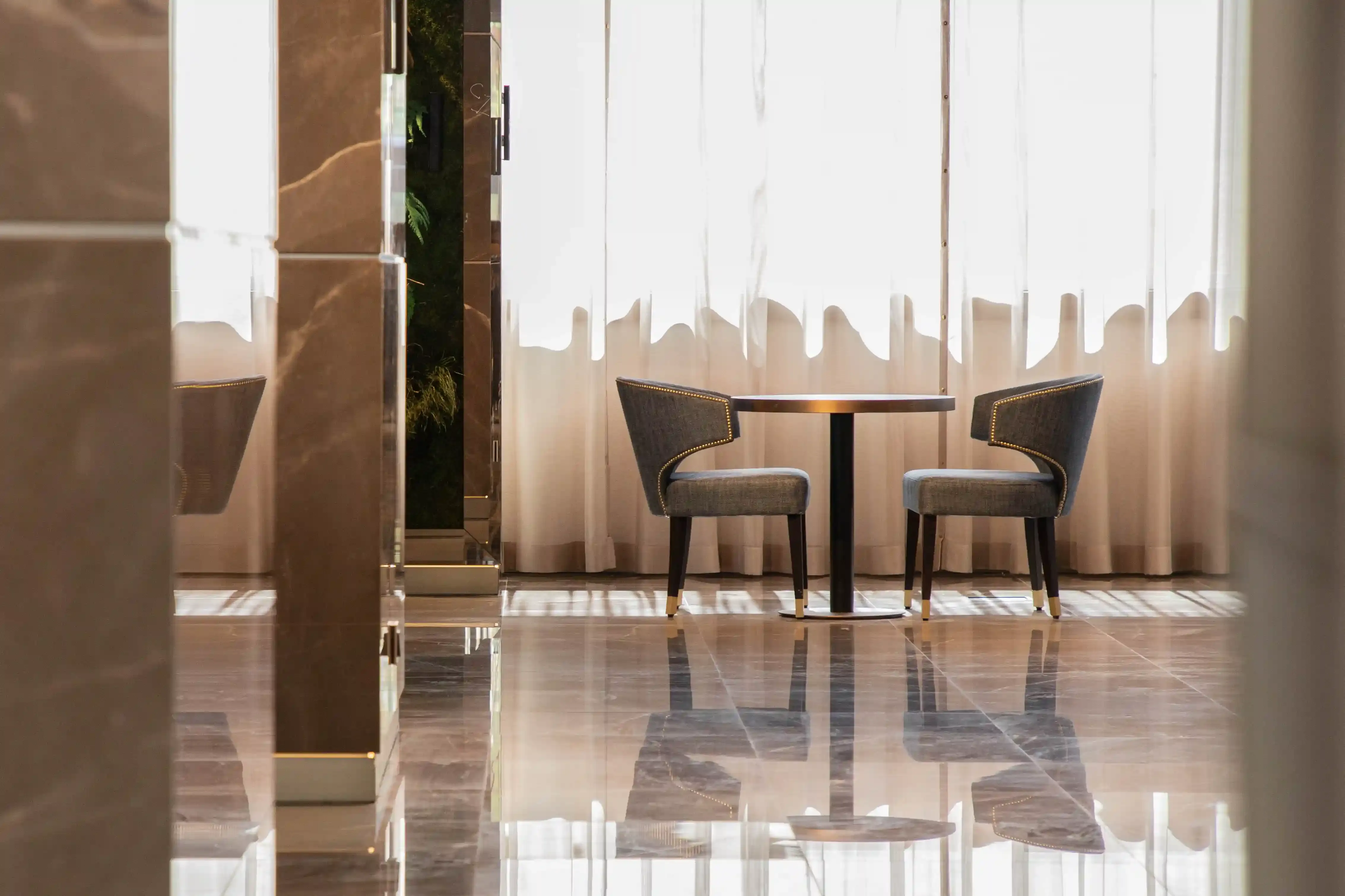 Mesa com duas cadeiras estofadas diante de cortinas claras, entre colunas de mármore, refletidas no pavimento polido.