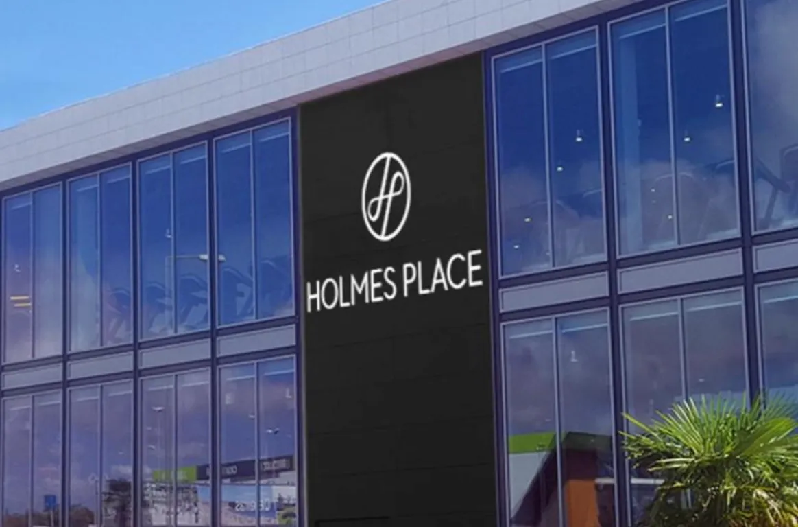 Fachada envidraçada de um ginásio com o logótipo e o nome Holmes Place num painel escuro ao centro e uma palmeira à direita.