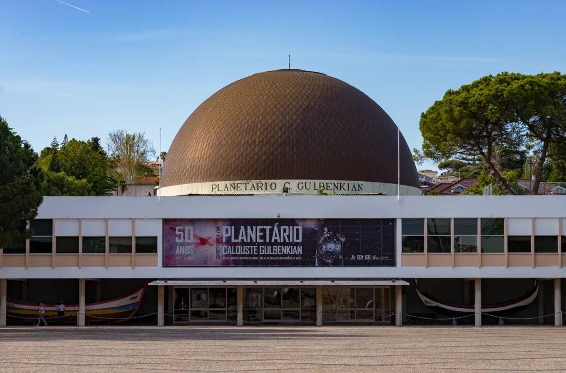 Cúpula acobreada sobre um edifício branco de dois pisos, com um painel do planetário na fachada, pinheiros e uma praça à frente.