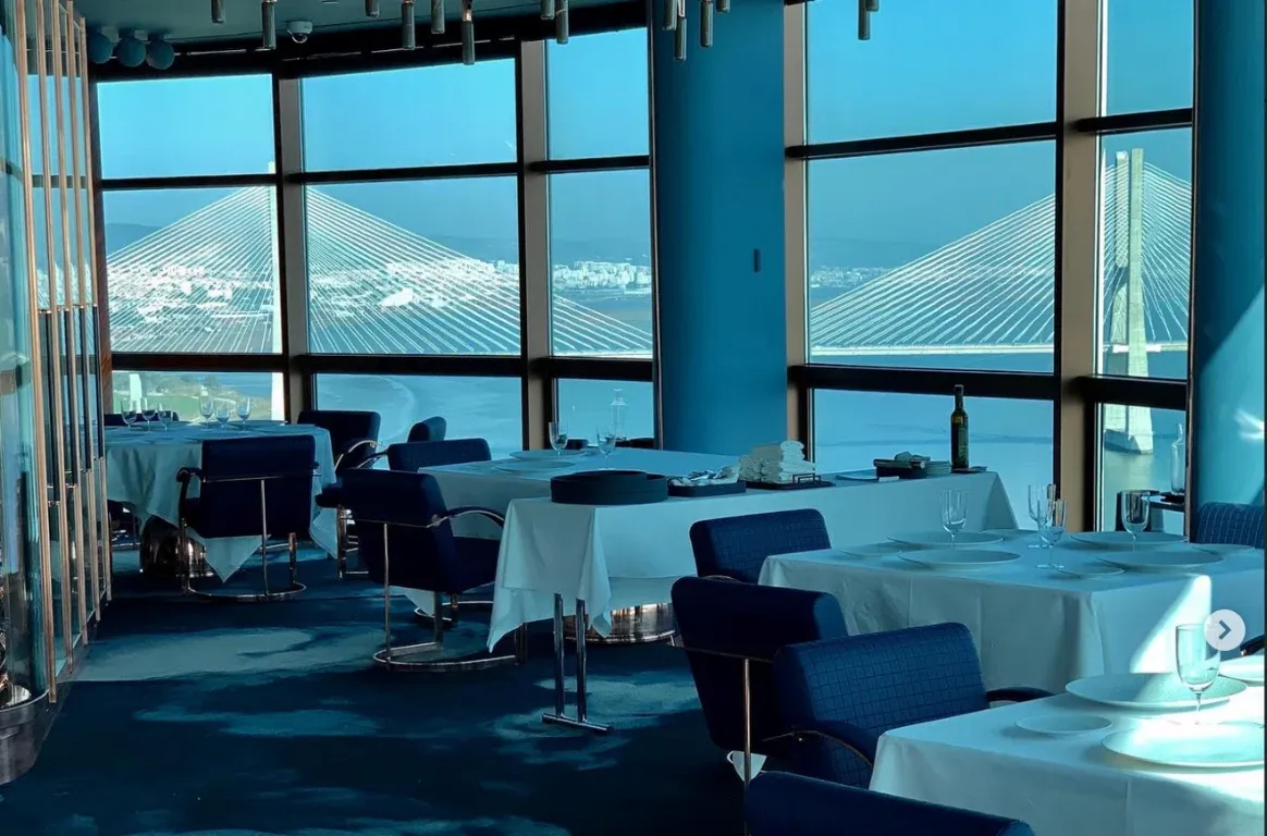 Sala de jantar com janelas amplas sobre o rio e uma ponte de tirantes, mesas de toalhas brancas e cadeiras azuis.