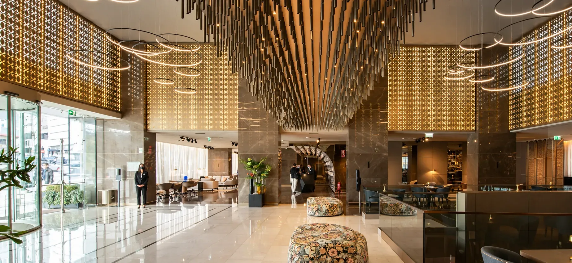 Átrio do hotel com teto de elementos verticais dourados, luminárias circulares suspensas e pavimento de mármore.