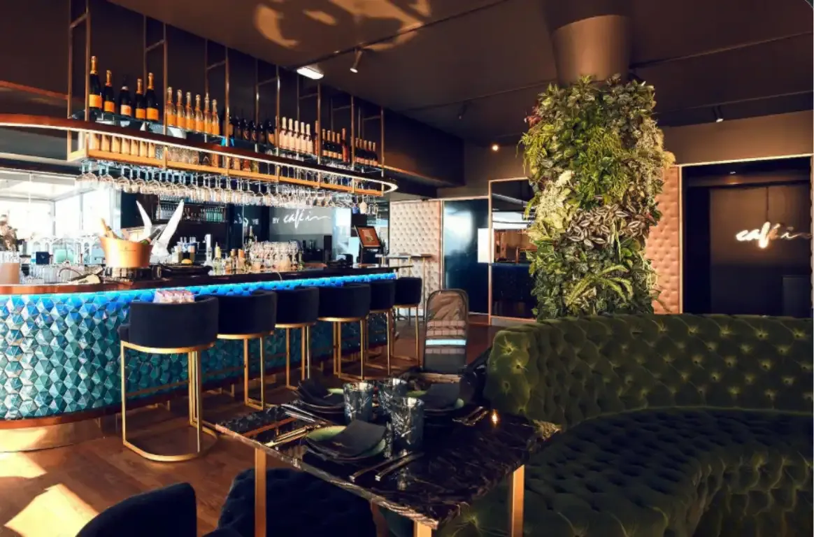 Bar com balcão em mosaico azul iluminado, bancos de veludo, prateleiras de garrafas e um sofá verde capitonê junto a uma coluna de plantas.