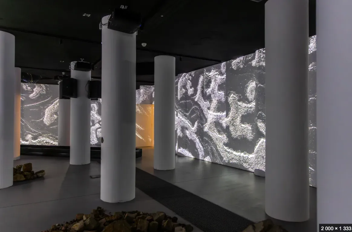 Sala escura com pilares brancos, projeções de texturas claras nas paredes, colunas de som suspensas e pedras soltas no chão.