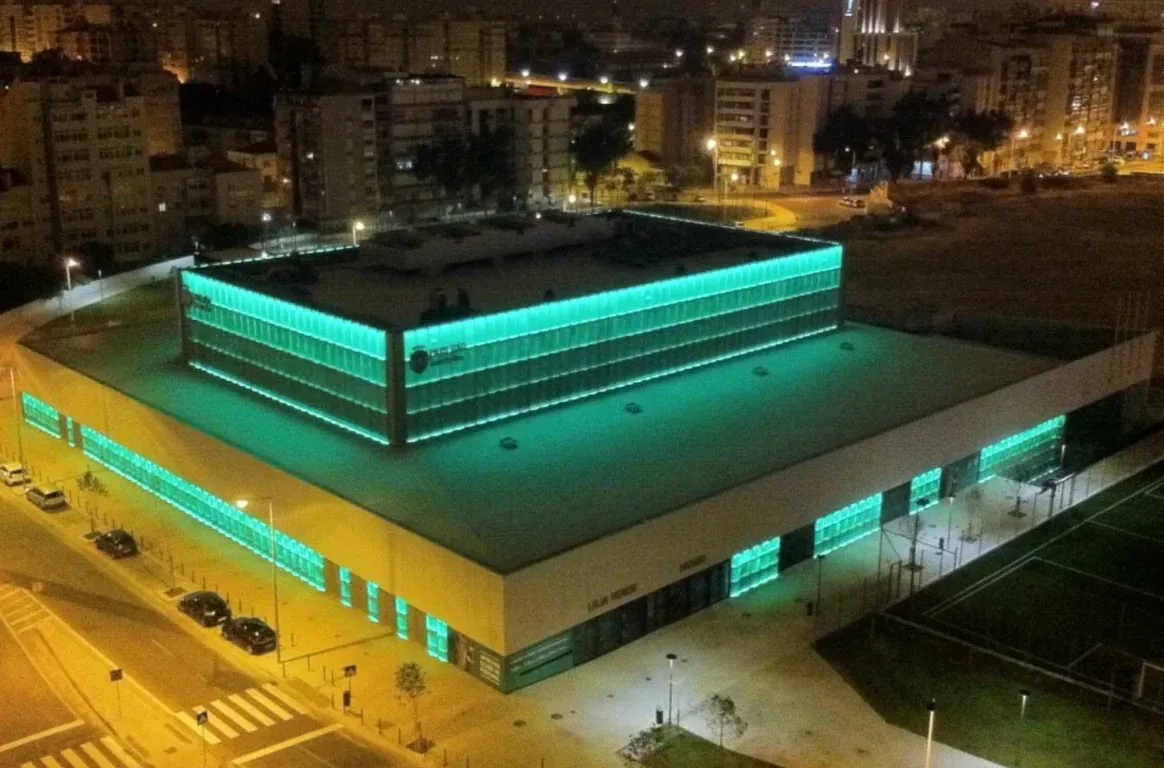 Pavilhão visto do alto, à noite, com as fachadas iluminadas a verde, a rua com carros estacionados e um campo de treino ao lado.