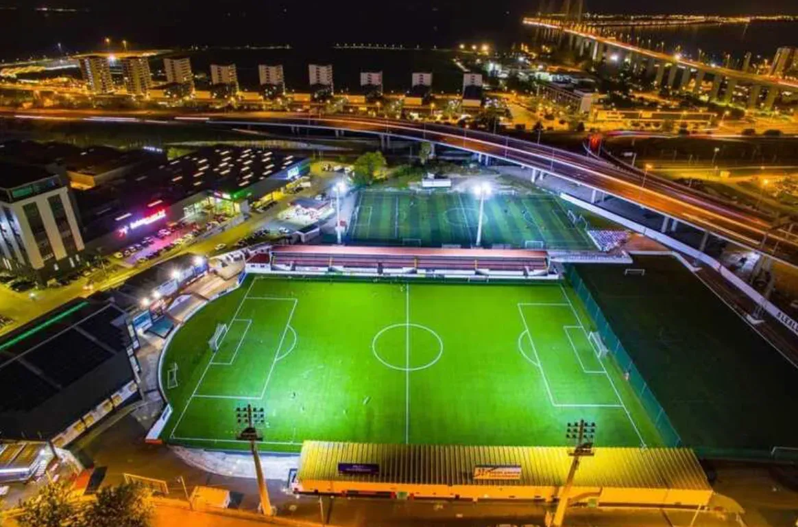 Vista aérea noturna de um campo de futebol relvado e iluminado, com a bancada coberta, outro campo atrás e uma ponte iluminada ao fundo.