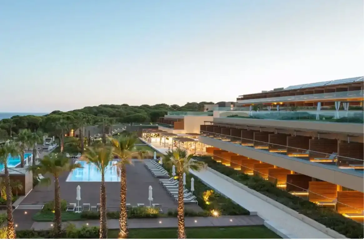 O hotel ao anoitecer, com terraços iluminados sobre um jardim de palmeiras e uma piscina.