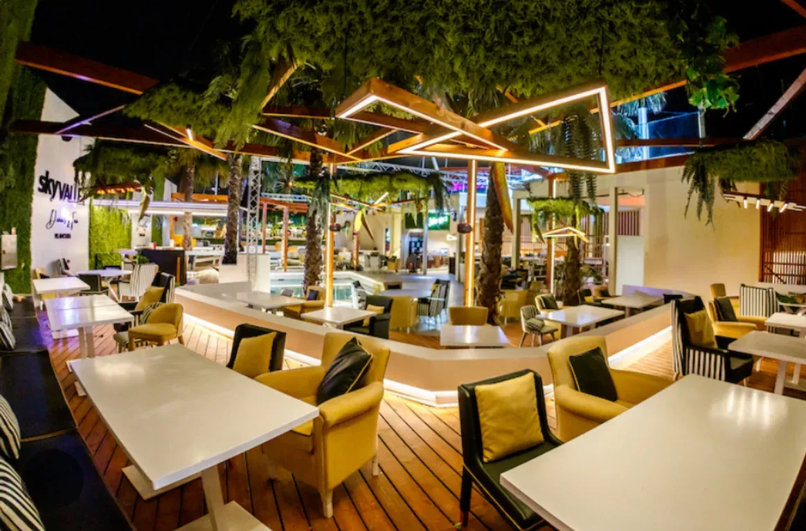 Esplanada à noite com mesas brancas e cadeirões amarelos sobre um deck de madeira, sob vegetação suspensa e molduras de luz LED.