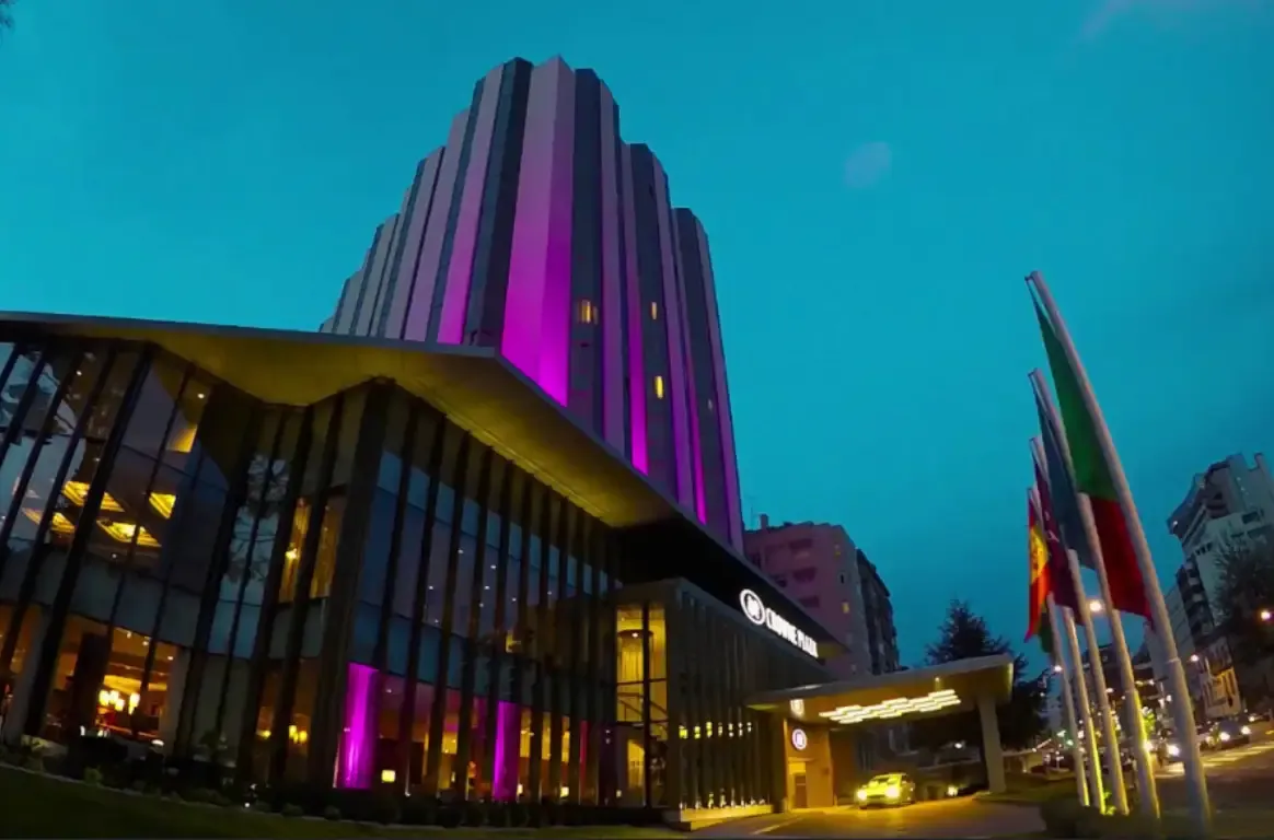 Fachada do hotel ao anoitecer, com a torre iluminada em magenta, a entrada envidraçada e bandeiras à frente.