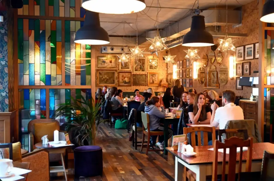 Sala cheia de clientes sob candeeiros industriais e estrelas de luz, com vitrais coloridos e quadros emoldurados nas paredes.