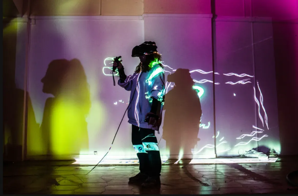 Pessoa com óculos de realidade virtual a desenhar no ar, diante de uma parede com traços de luz projetados e sombras coloridas.