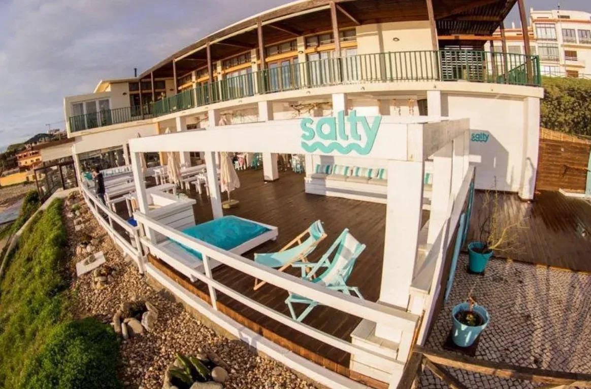 Esplanada de deck de madeira com o letreiro salty, espreguiçadeiras e uma cama de praia azul-turquesa, sobre uma encosta de seixos.