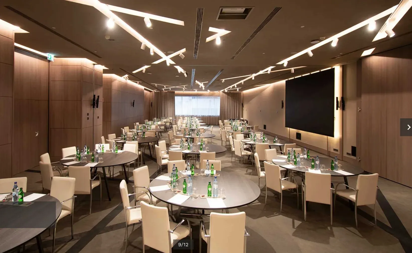 Sala de eventos montada com mesas redondas, iluminação linear no teto, um ecrã ao fundo e outro na parede lateral.