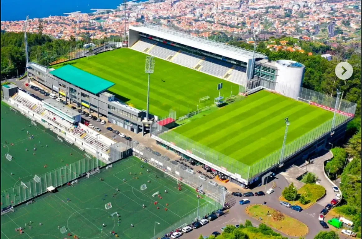 Vista aérea de um estádio de relvado verde na encosta, com a bancada coberta, campos de treino ao lado e casas da cidade em fundo.
