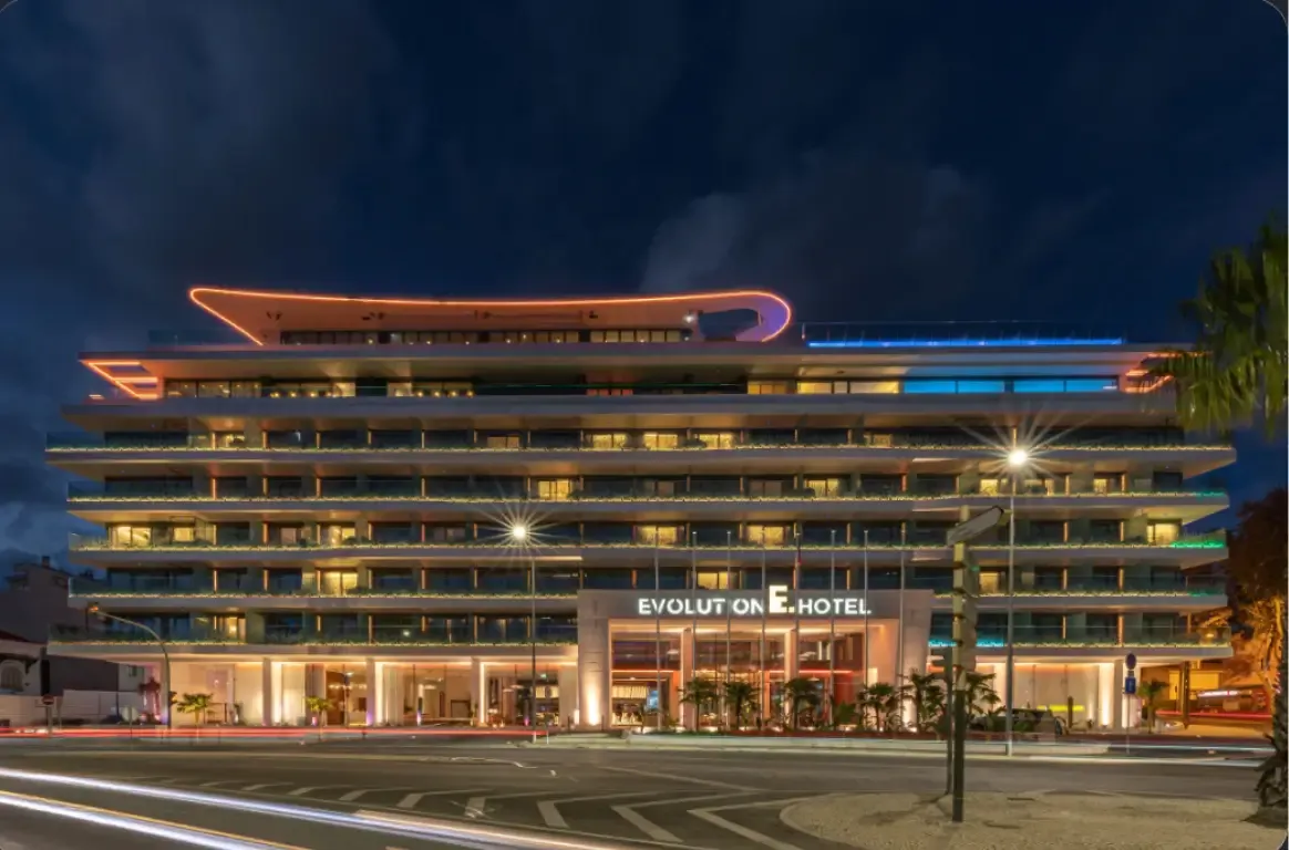 Fachada do hotel à noite, com varandas a toda a largura, letreiro iluminado e linhas de luz na cobertura.