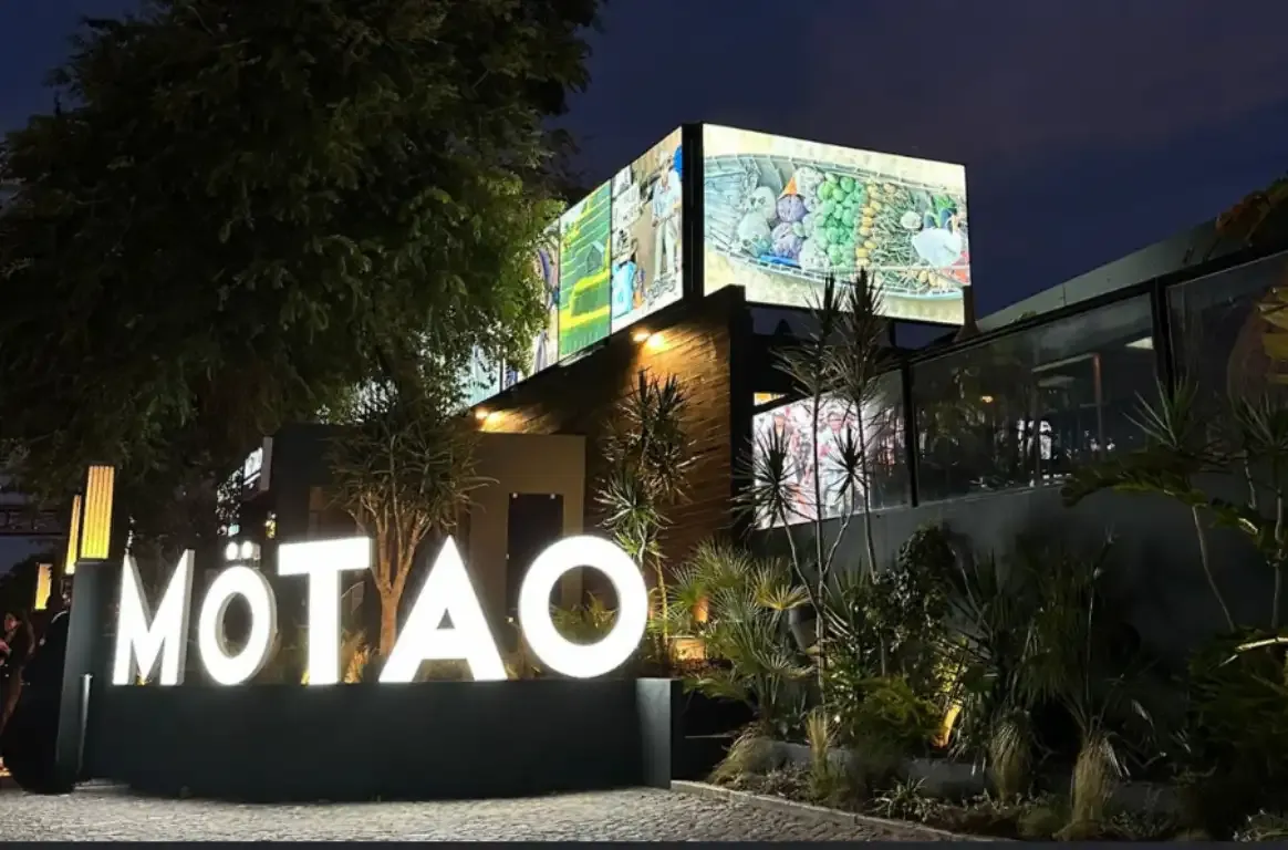 Letreiro luminoso MOTAO à noite, à entrada de um edifício com ecrãs na fachada e um jardim de plantas tropicais.