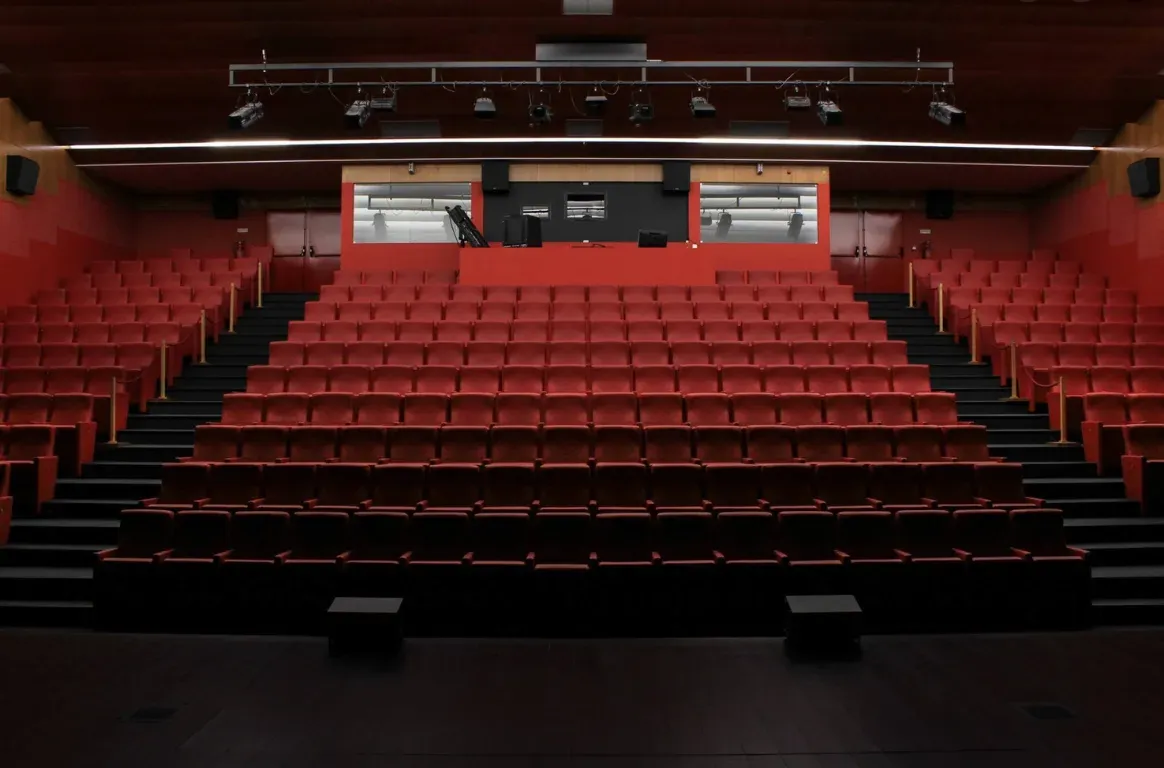 Auditório de cadeiras vermelhas em bancada, visto do palco, com uma barra de focos de luz no teto e a cabine técnica ao fundo.