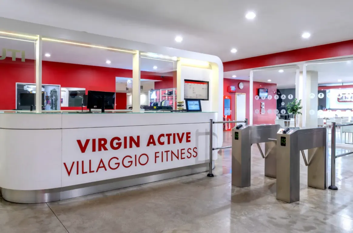 Receção de um clube Virgin Active, com balcão branco de letras vermelhas, paredes vermelhas e torniquetes de acesso em inox.