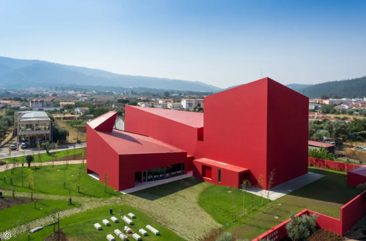 Edifício vermelho de volumes angulosos, visto de cima, rodeado de relvados, com casas e montanhas ao fundo.