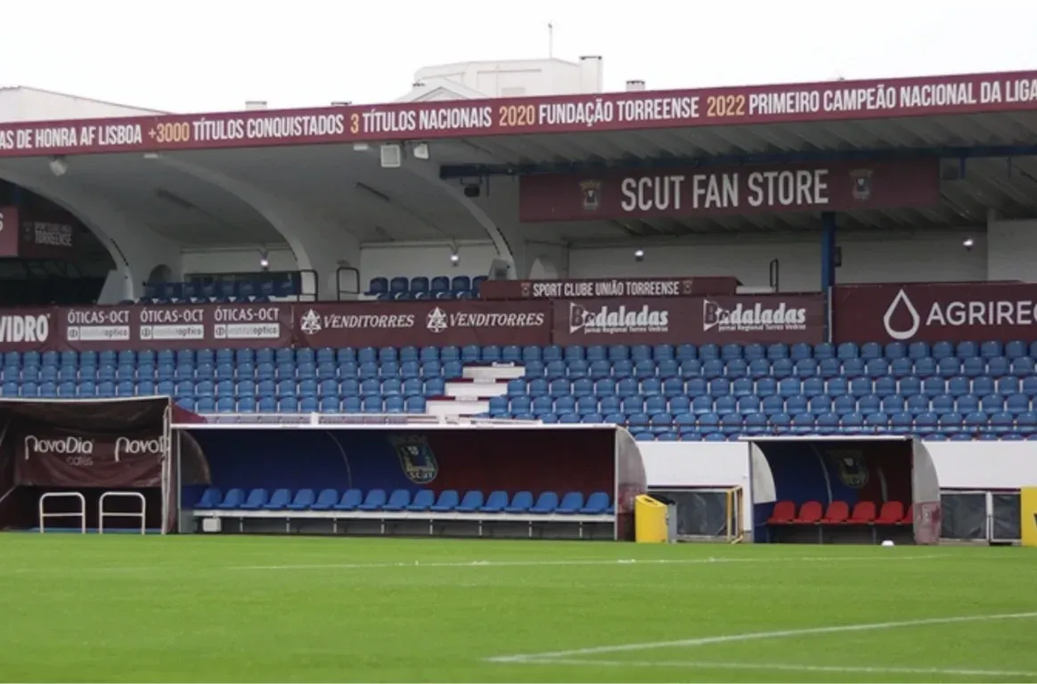 Bancada de cadeiras azuis com faixas do Sport Clube União Torreense, bancos de suplentes e o relvado em primeiro plano.
