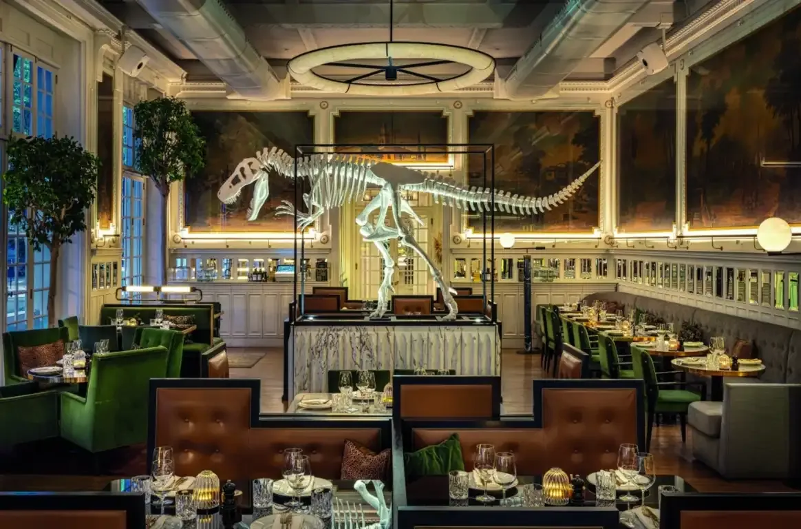 Sala com um esqueleto de dinossauro numa vitrina ao centro, sofás de couro, cadeirões verdes e grandes pinturas nas paredes.