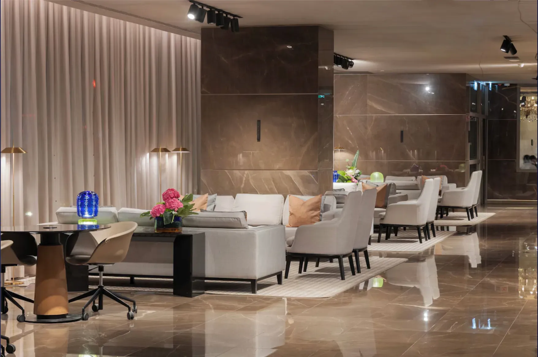 Lounge com sofás e poltronas claras, cortinas e pavimento de mármore.