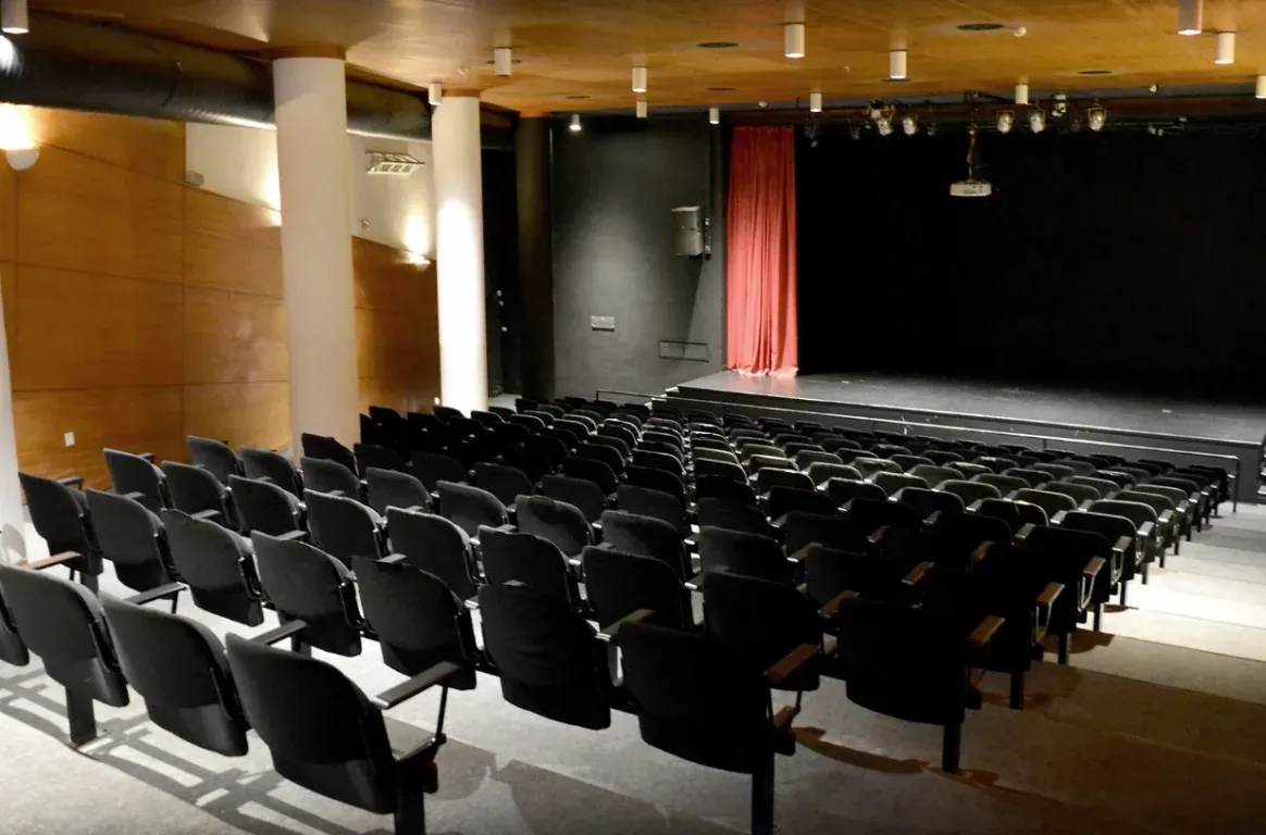 Pequeno auditório de cadeiras pretas virado para um palco escuro, com uma cortina vermelha, focos de luz no teto e paredes de madeira.