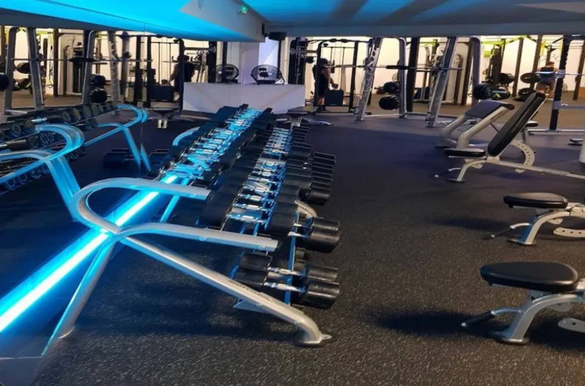 Sala de musculação com halteres alinhados em suportes iluminados a azul, bancos de treino e máquinas ao fundo.