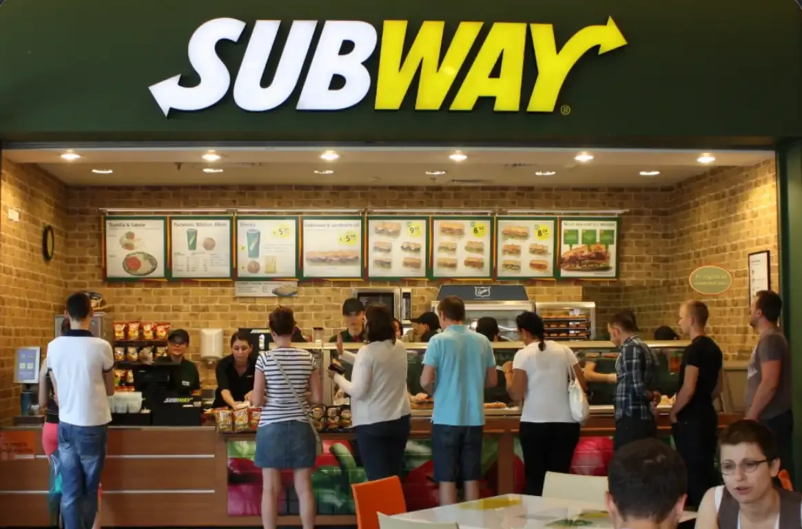 Clientes em fila diante do balcão de um restaurante Subway, com o letreiro da marca e os painéis do menu iluminados por cima.