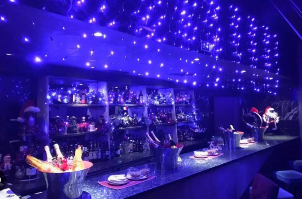Balcão de bar iluminado a azul, com cortinas de pequenas luzes, prateleiras de garrafas e baldes de champanhe sobre o tampo.
