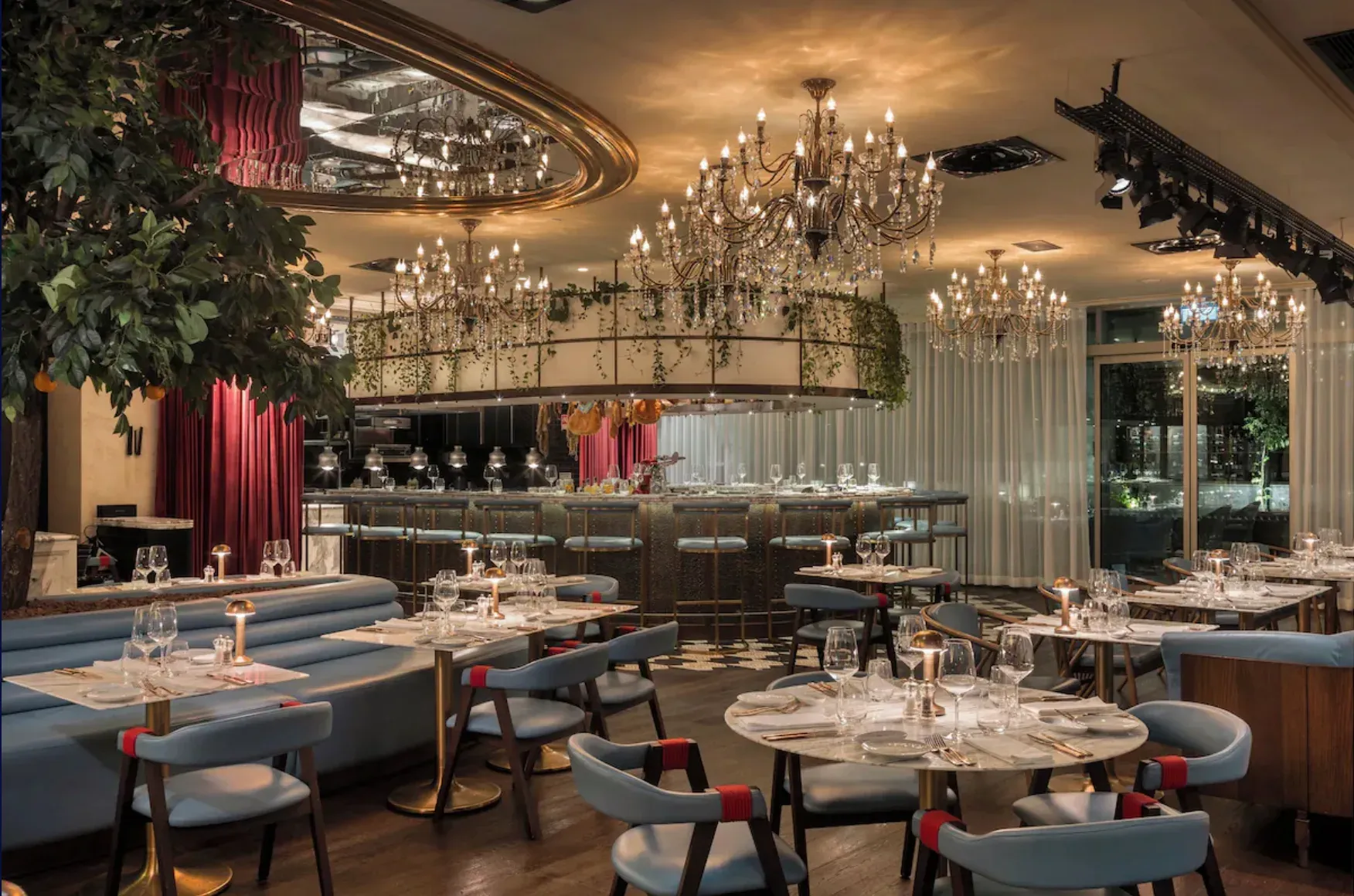 Restaurante com lustres de cristal, bancos azuis, mesas postas e um bar ao fundo.