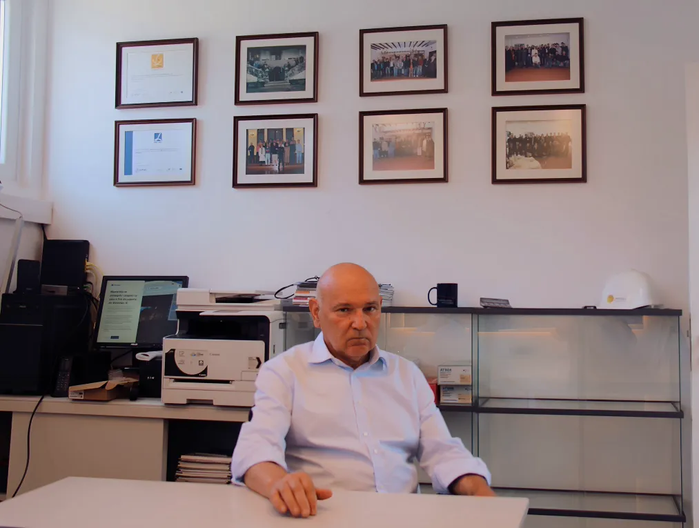 Vítor Alves à mesa no mesmo escritório, num plano mais aberto, com equipamento de escritório e uma vitrine de vidro por trás.