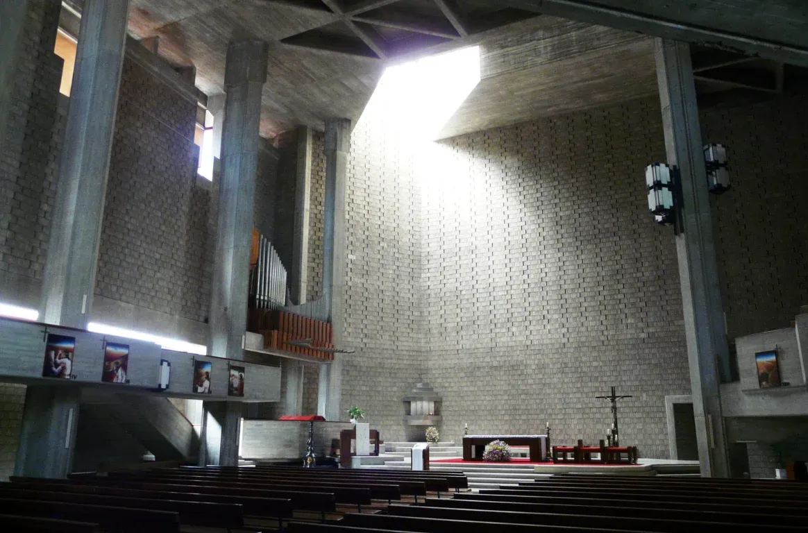 Interior de betão e tijolo com luz zenital sobre o altar, um crucifixo, o órgão num balcão lateral e filas de bancos de madeira.